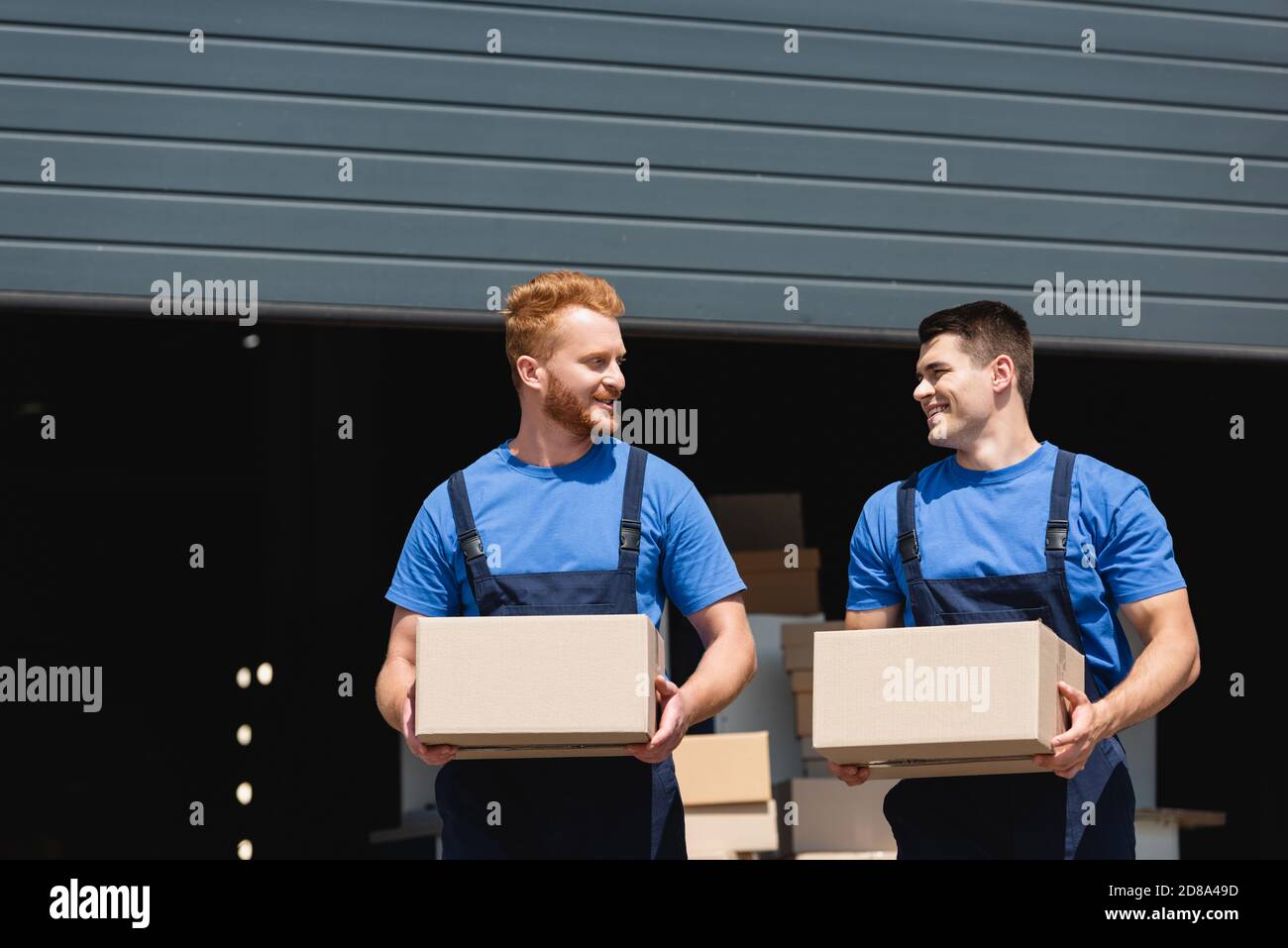 Young movers in uniform looking at each other while holding cardboard boxes on urban street Stock Photo
