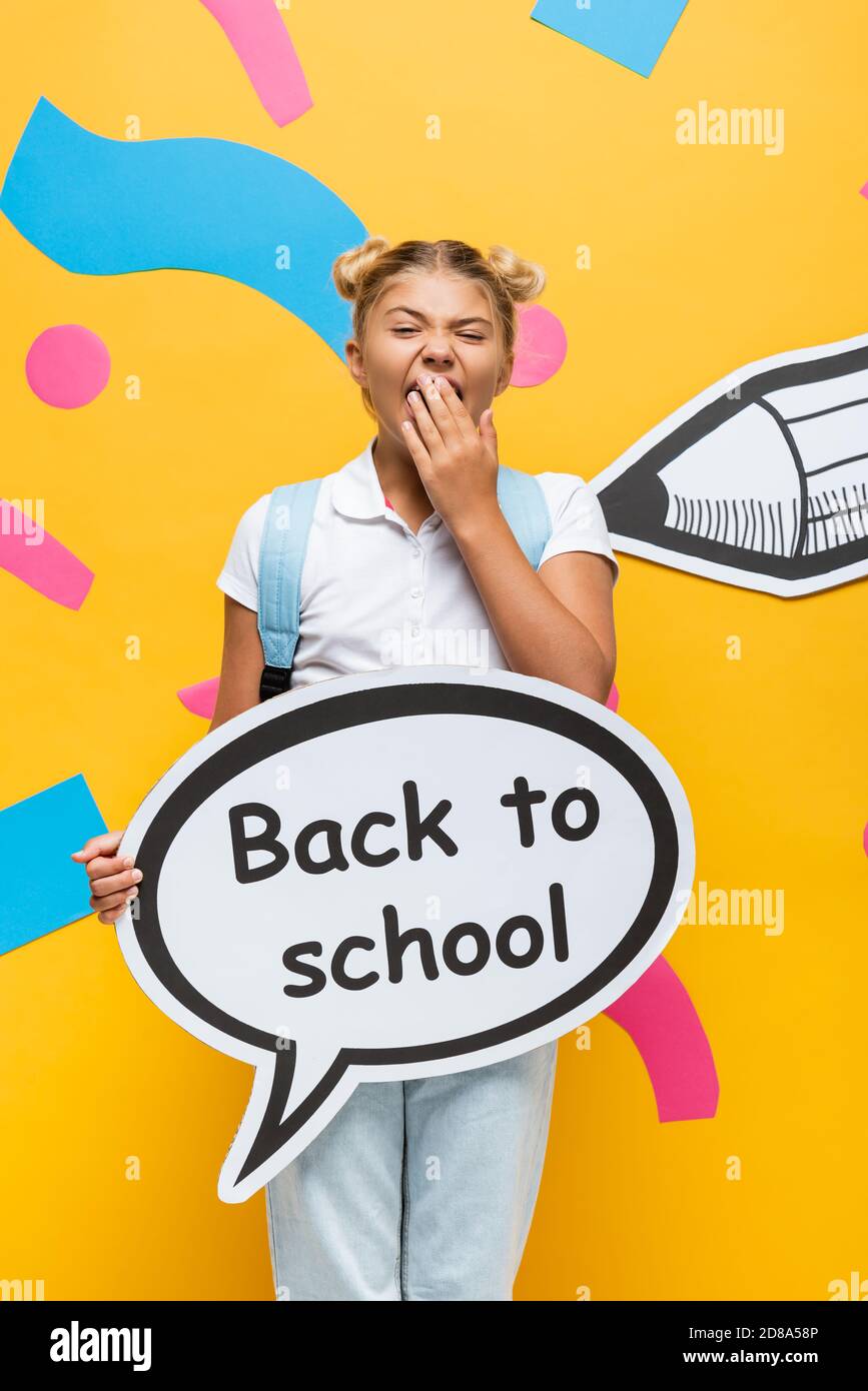 bored schoolgirl yawning while holding speech bubble with back to school lettering near paper pencil and colorful elements on yellow Stock Photo