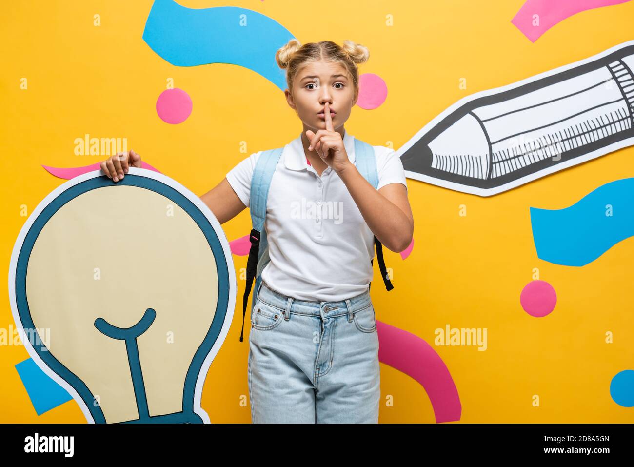 Schoolgirl showing quiet gesture while holding decorative light bulb near paper art on yellow background Stock Photo