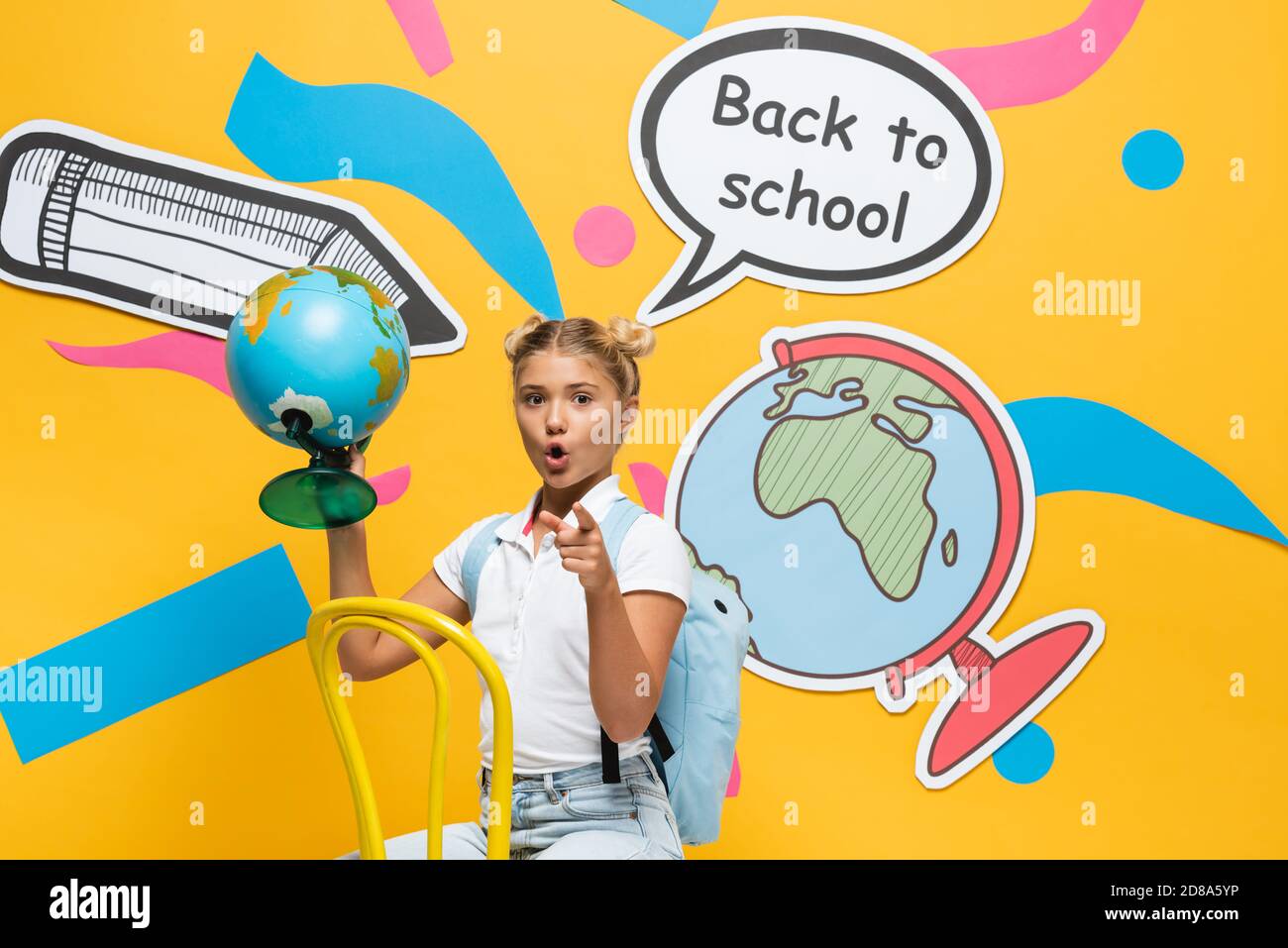 Excited schoolgirl pointing at camera while holding globe near paper art on yellow background Stock Photo