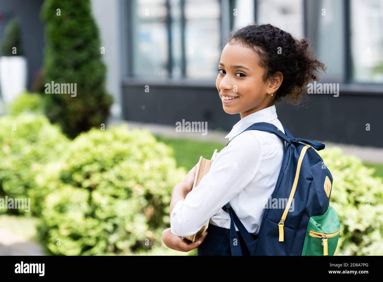 side view of smiling african american schoolgirl with book outdoors ...