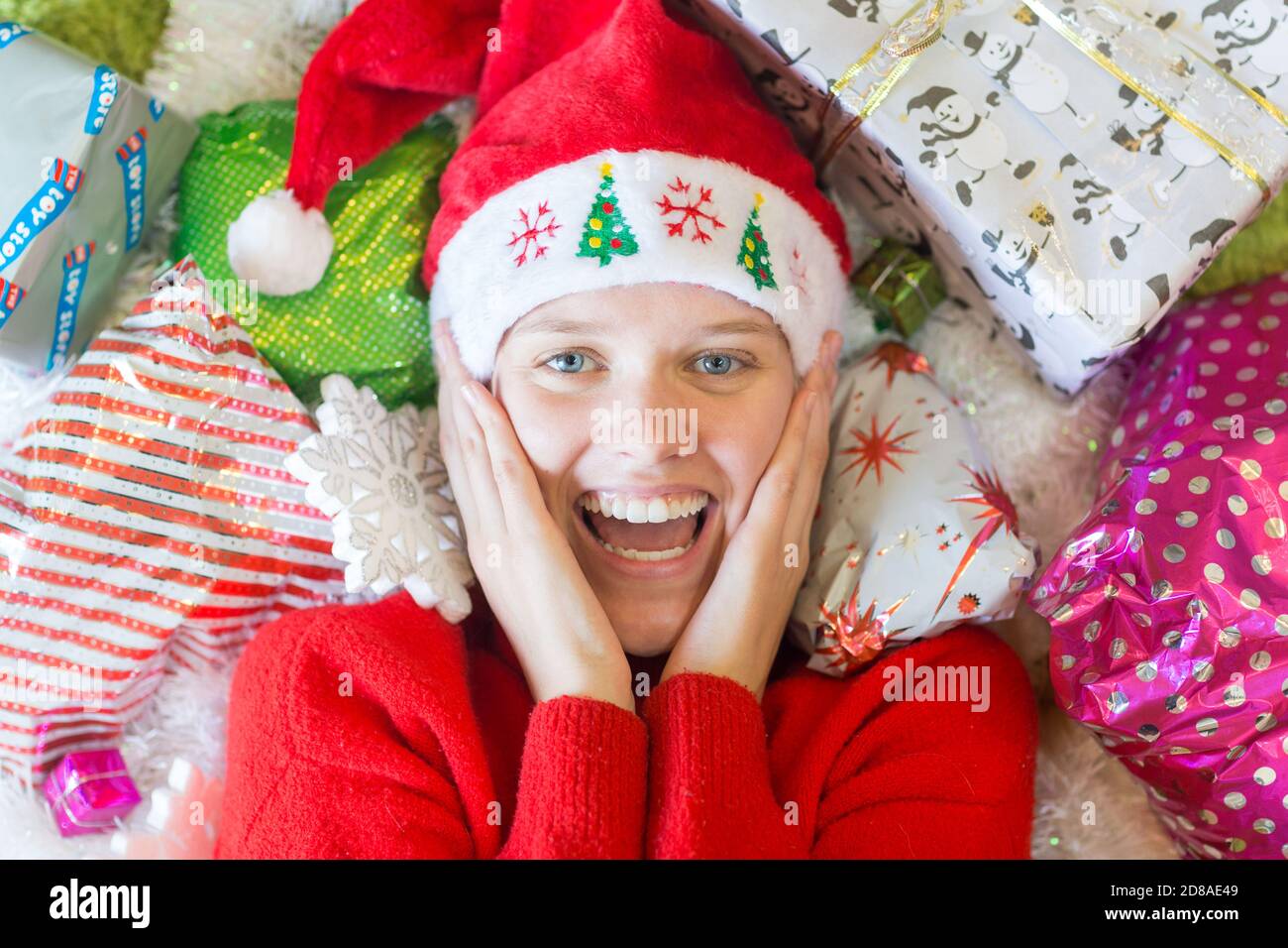 Christmas time! Joyful excited young woman lying surrounded by many gifts wearing a red Santa hat. Stock Photo