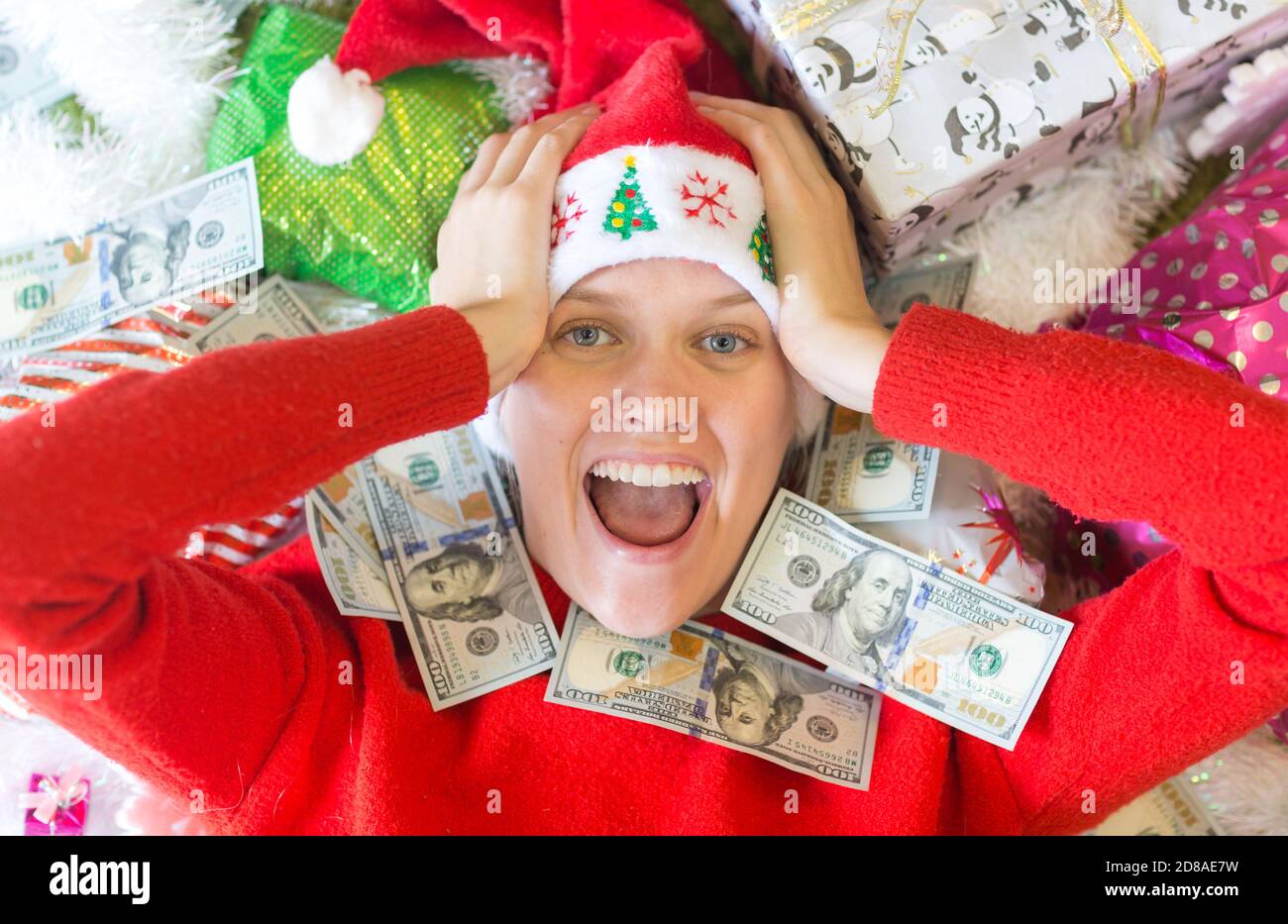 Amazed excited young woman surrounded by cash money bills and lots of Christmas gifts. Stock Photo