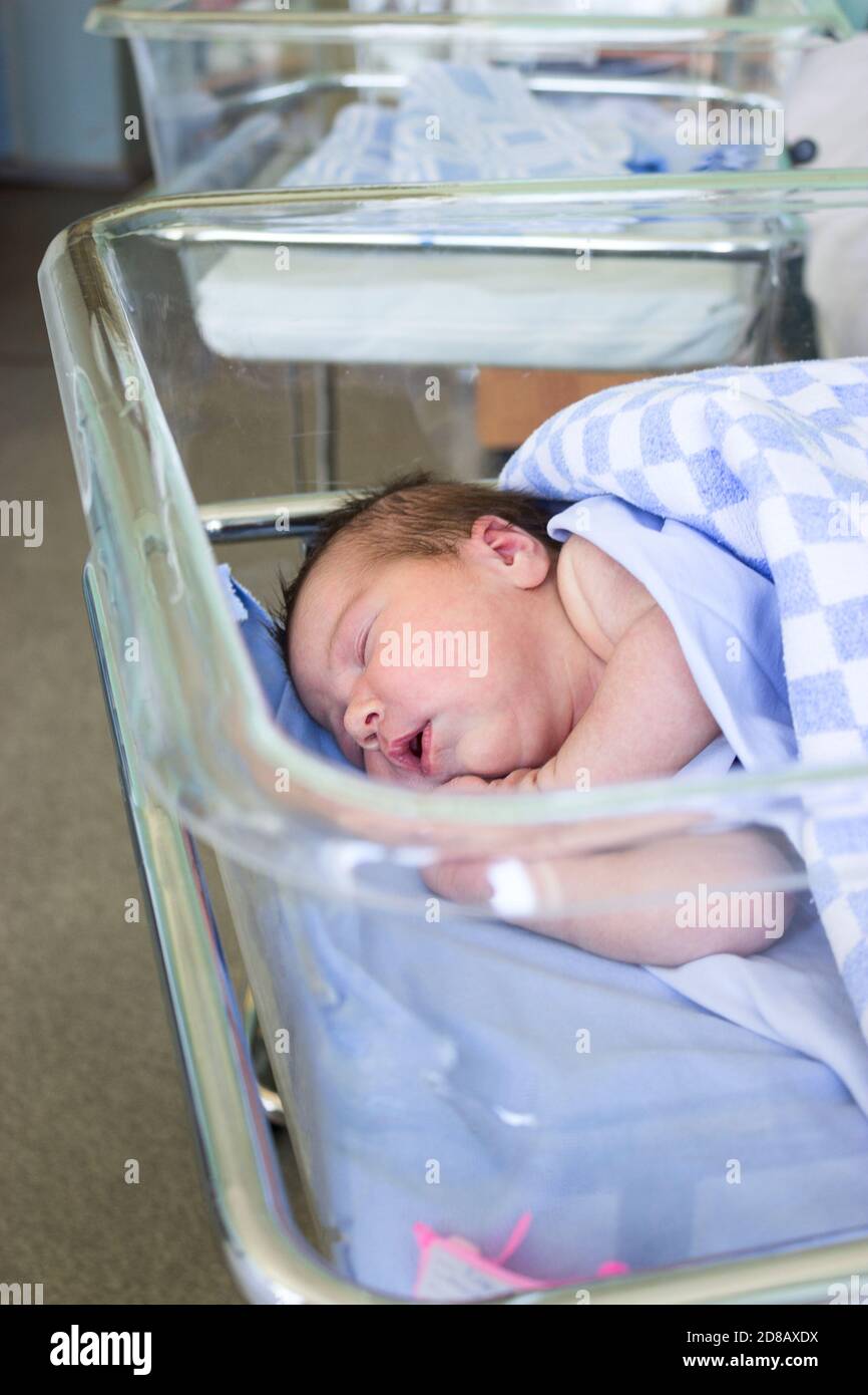 Cute Caucasian little newborn baby sleeping in hospital bed, folded hands with open mouth, lying under blanket Stock Photo