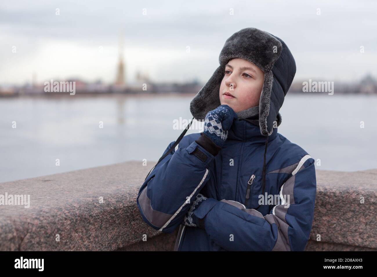 Teenager dressed warm jacket and hat with earflaps thinking, Peter and Paul fortress is on background, granite bridge across Neva river in St. Petersb Stock Photo
