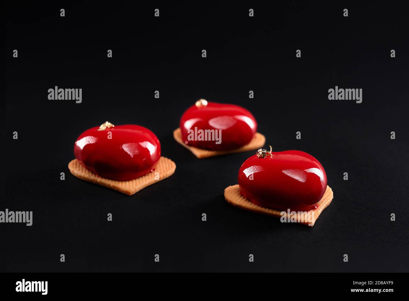 Red heart cakes decorated with gold isolated on black background. Dessert with smooth surface, mousse and mirror glaze on sweet cookies. Delicious dish in cafeteria. Stock Photo