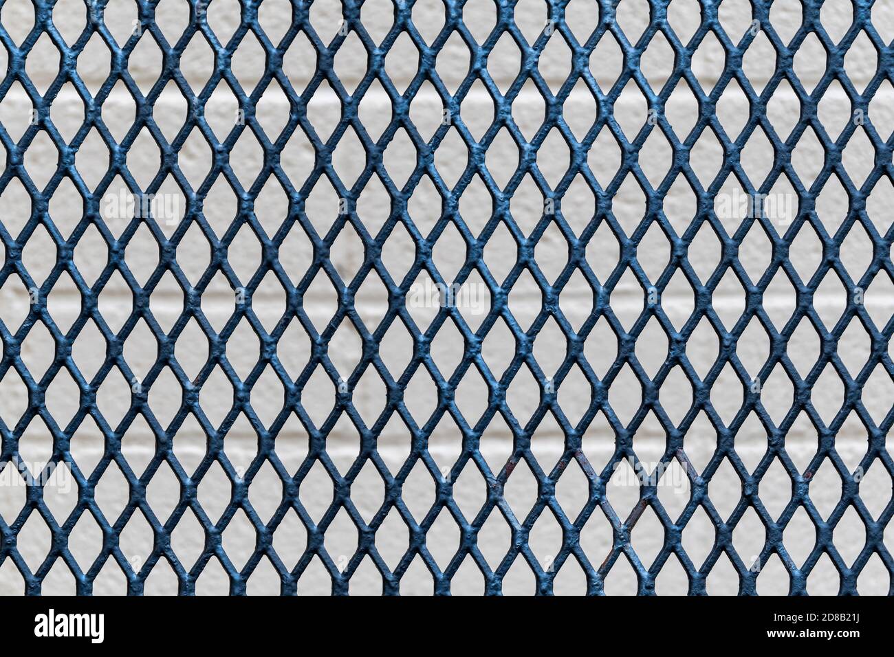 A diamond grid pattern in a metal fence. Cinder block wall behind, outside depth of field. Stock Photo