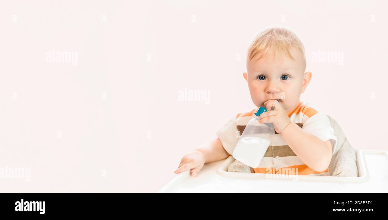 A small happy blue-eyed child is sitting at the table and drinking water from a bottle with a pacifier. Independent. Copy space Stock Photo