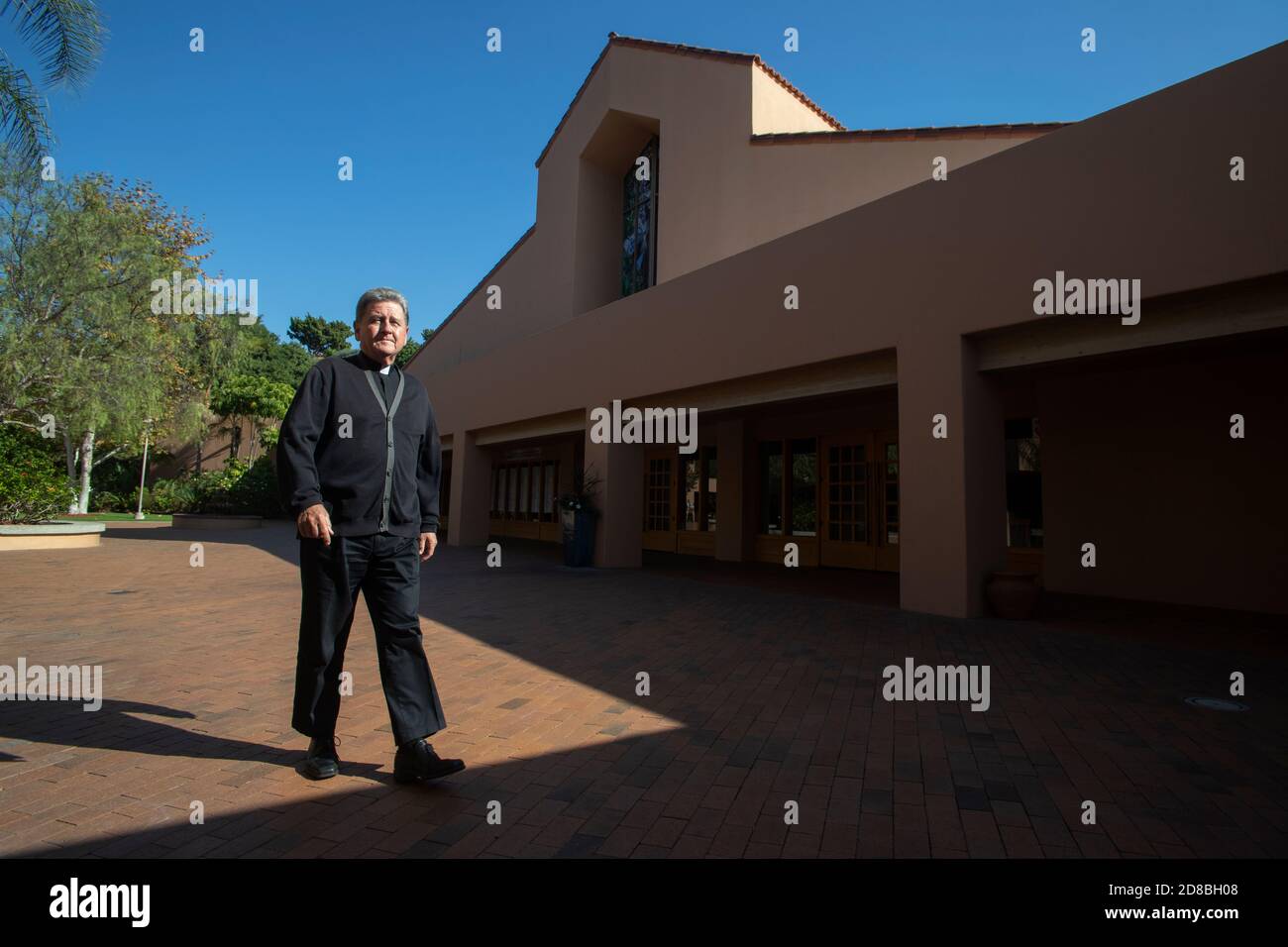 The monsignor of a Southern California Catholic church walks across the church courtyard in after sun and shadow. Stock Photo