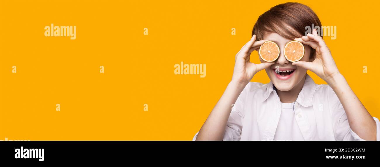 Smiling caucasian boy is covering his eye with sliced lemon pieces posing on a yellow wall with free space Stock Photo