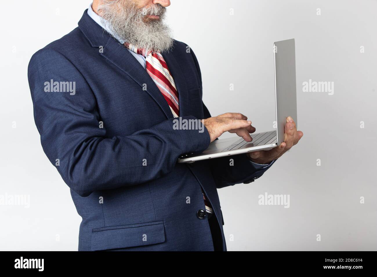 Portrait of a 60-65 year old man with a suit and a beard surfing the internet on a laptop. Lifestyle concept, technology. Isolated on white background Stock Photo