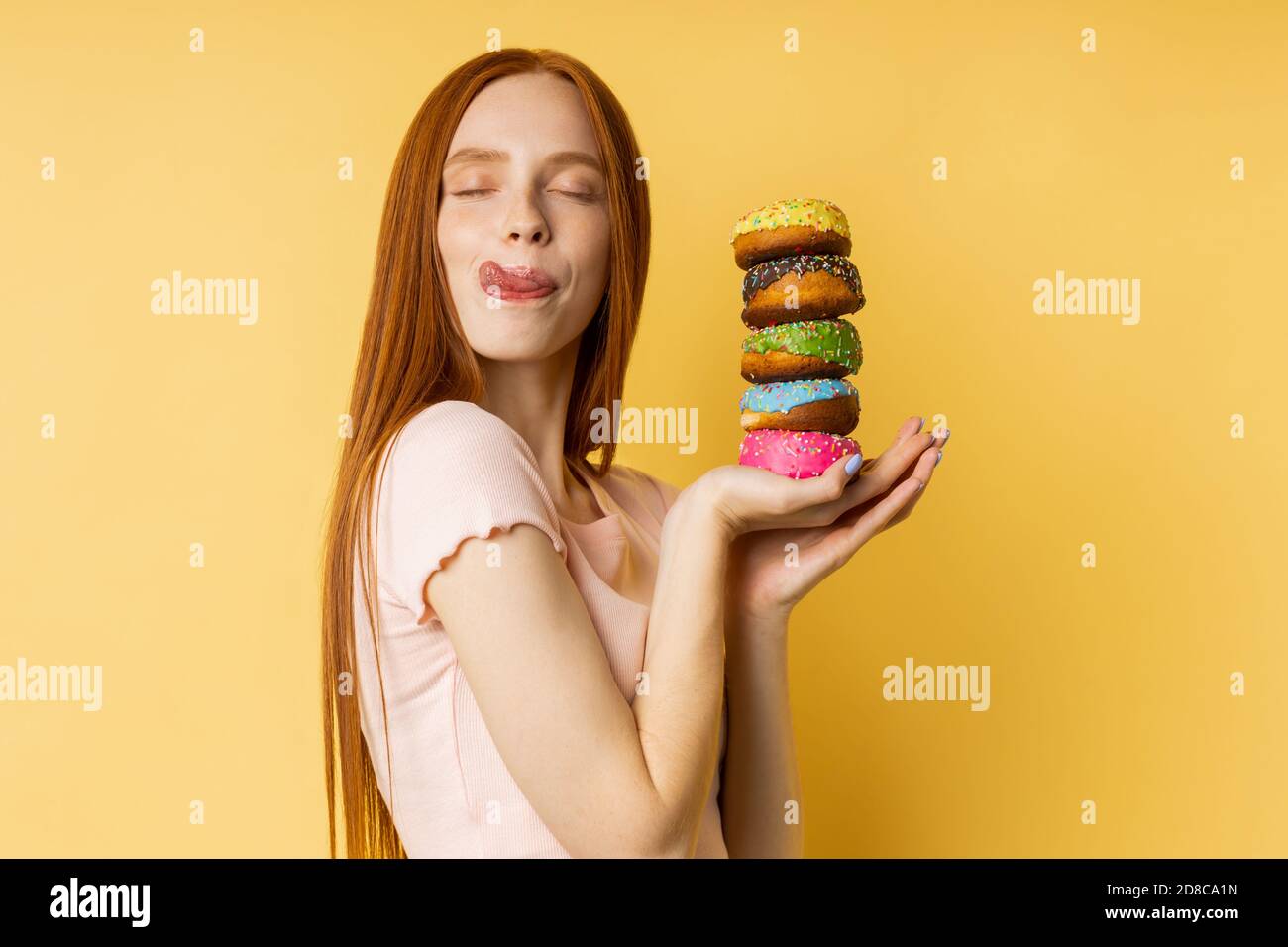 Shot of happy caucasian woman with red hair, closed eyes, licking her lips with tongue, holding in hands delicious colorful donuts isolated over yello Stock Photo