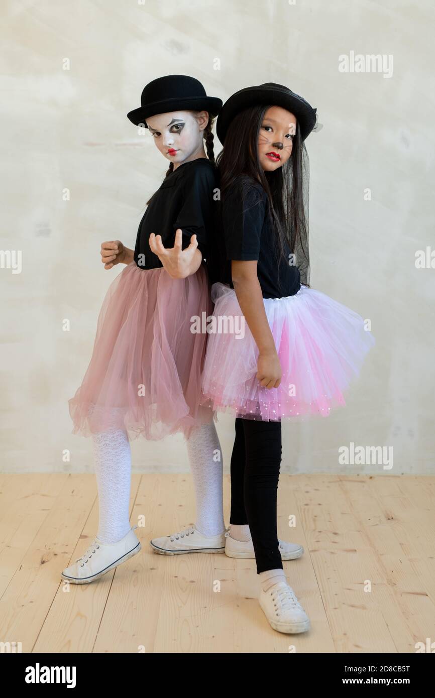 Portrait of serious multi-ethnic girls in costumes consisting of black sweaters and pink tutu skirts posing together against wall Stock Photo