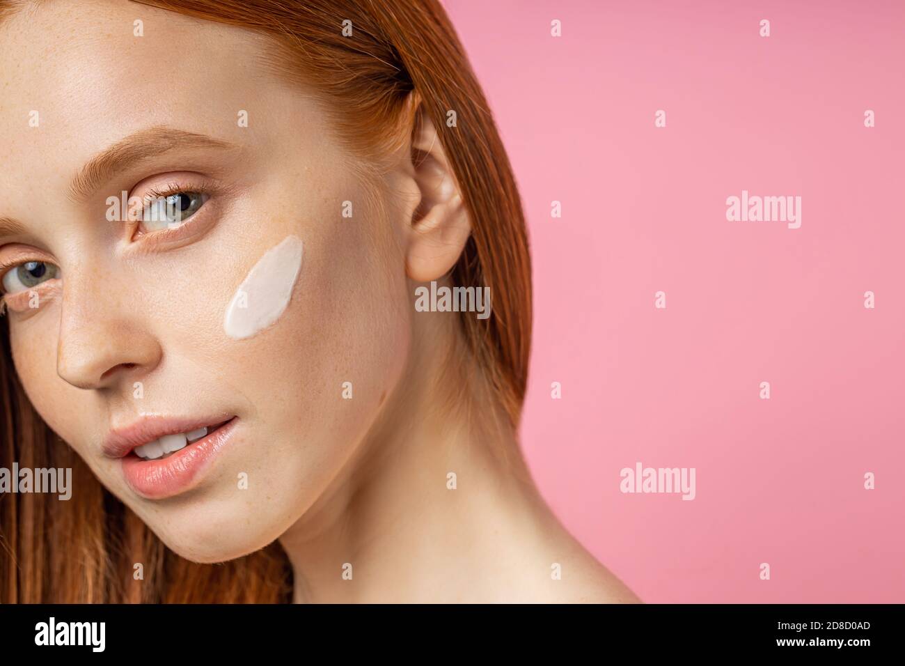 Cropped close up portrait of young woman with cream on cheek. Charming caucasian girl with red hair, fresh freckled skin, smiling happily standing aga Stock Photo