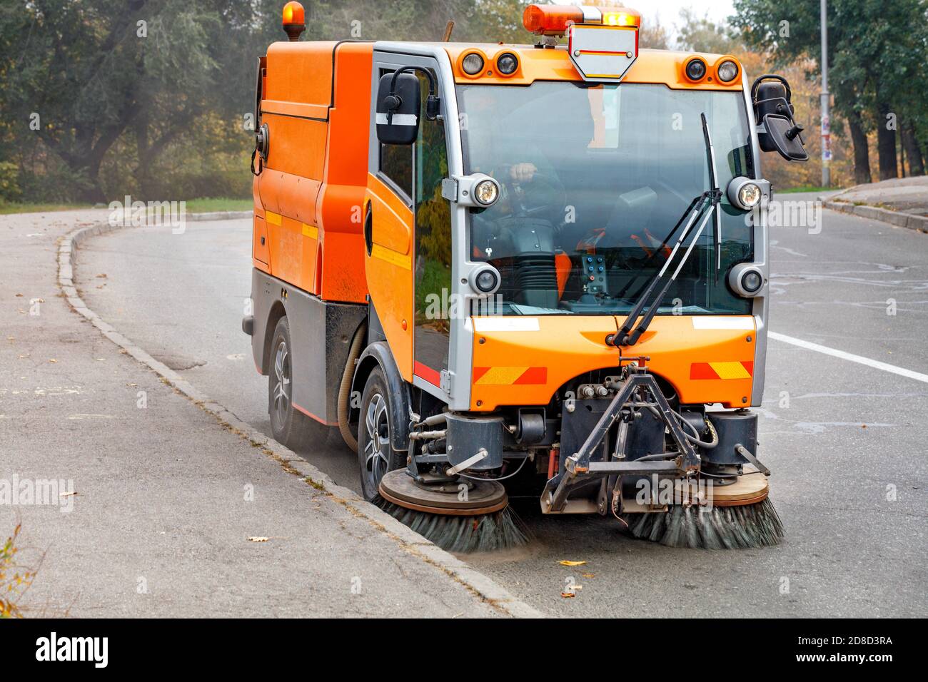 A small street sweeper with two independently controllable, hydraulically adjustable front brushes cleans the road in a residential area of the city. Stock Photo