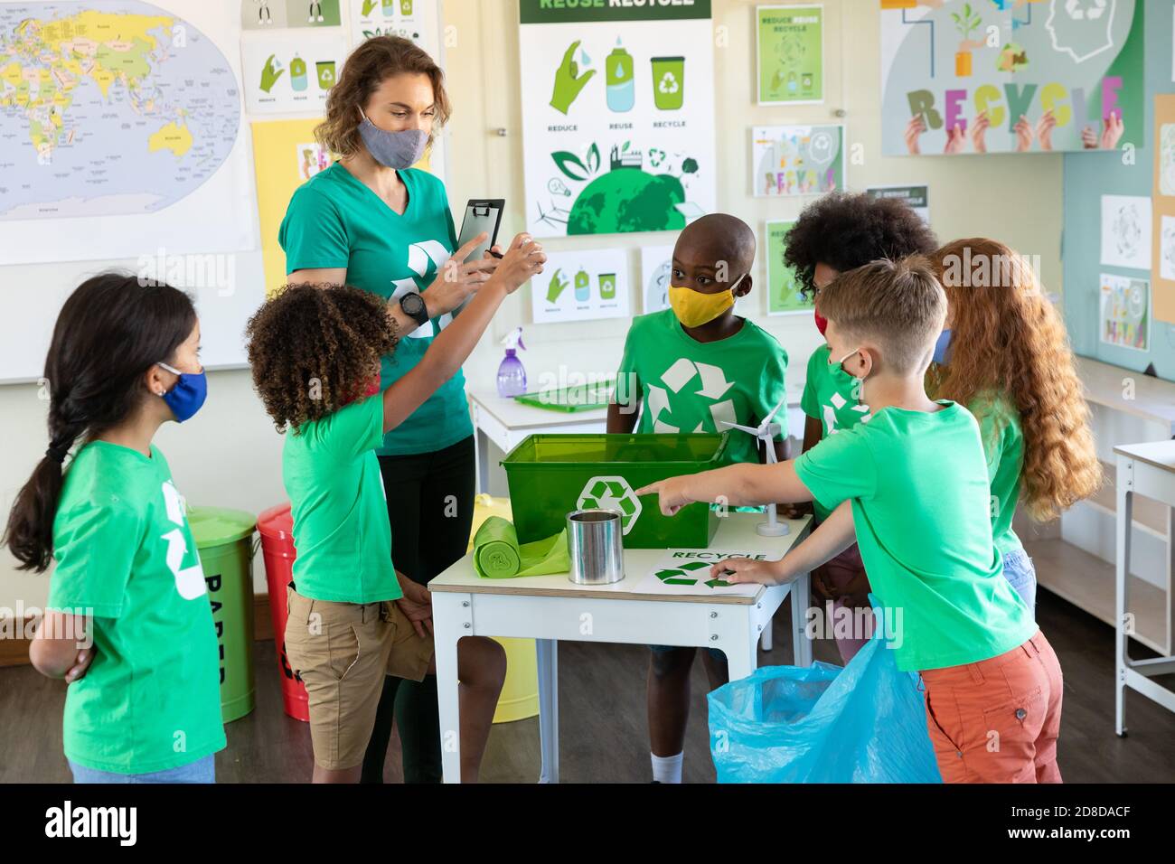 Female teacher and students wearing face masks with recycling container on table in class Stock Photo