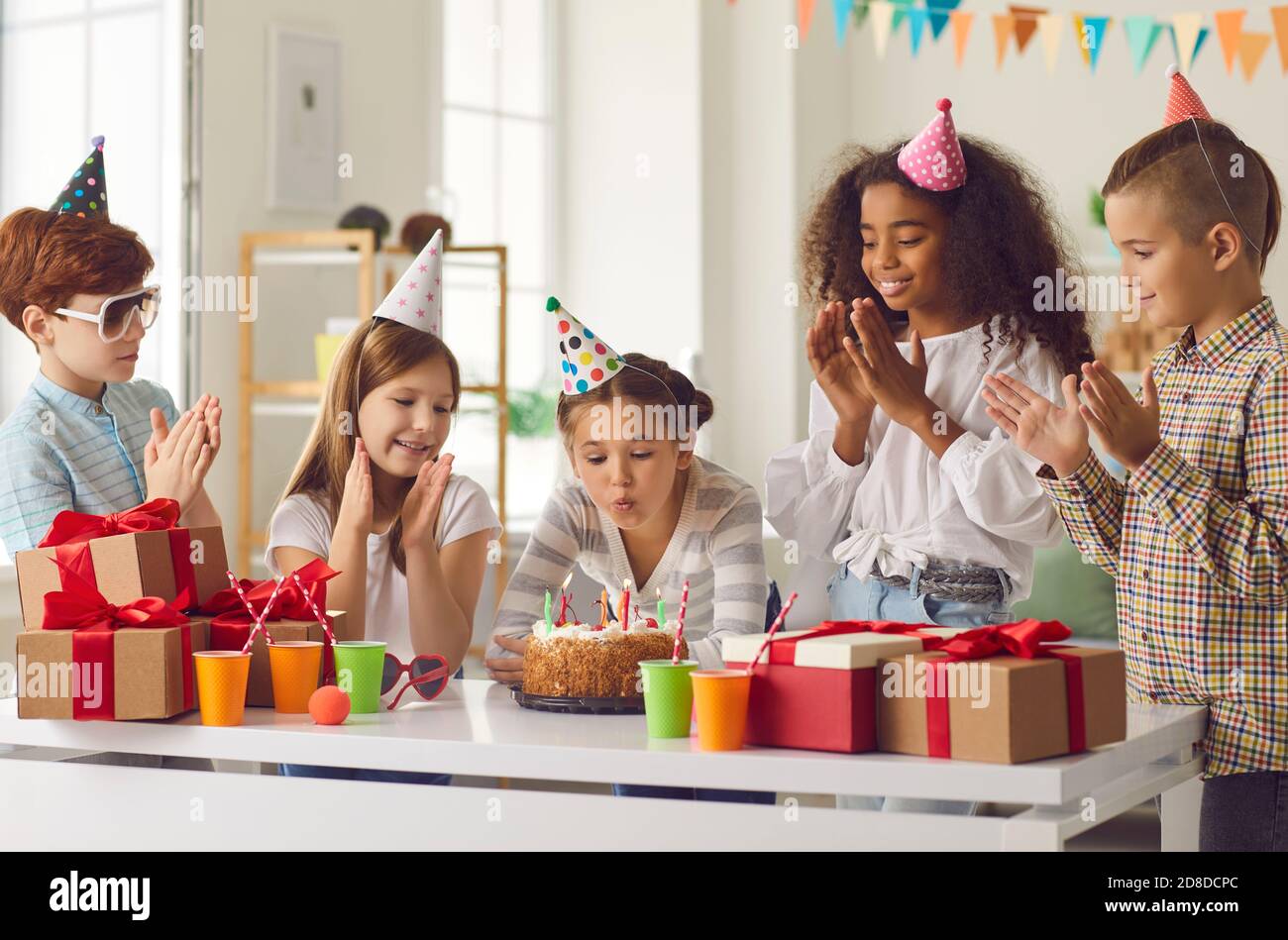 Little birthday girl blows out candles on a cake to the applause of her international friends. Stock Photo