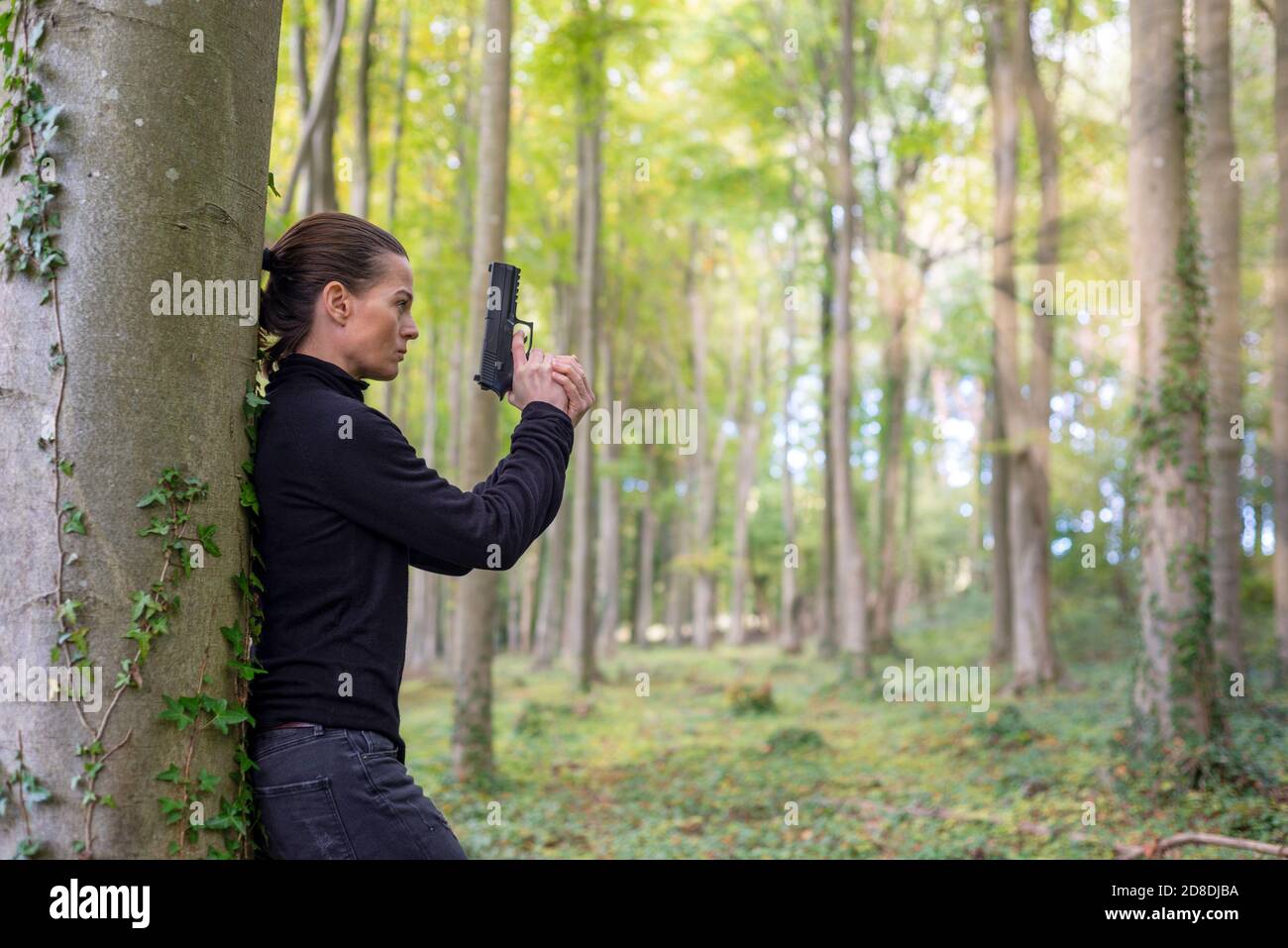 Woman holding a pistol, with her back against a tree. Stock Photo