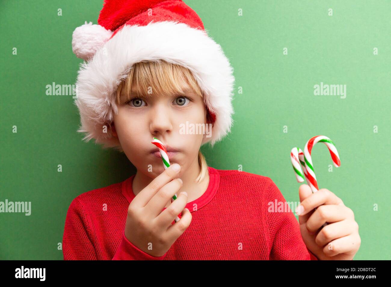 Boy in Santa Claus hat and a red jacket, eating candy canes on a green background. Christmas concept Stock Photo
