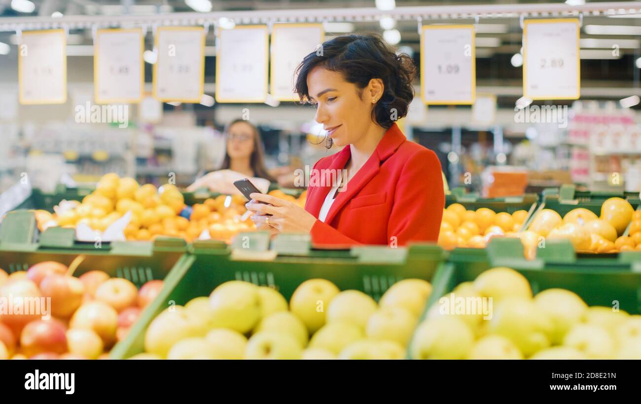 At the Supermarket: Portrait of the Beautiful Woman Using Smartphone, Chooses Products In the Fresh Produce Aisle. Woman Immersed in Internet Surfing Stock Photo