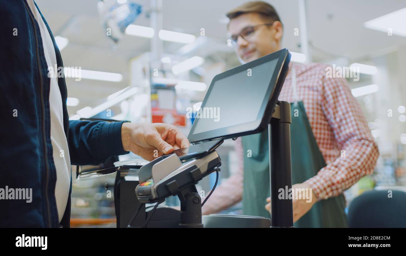 At the Supermarket: Checkout Counter Customer Pays with Smartphone for His Food Items. Big Shopping Mall with Friendly Cashier, Small Lines and Modern Stock Photo