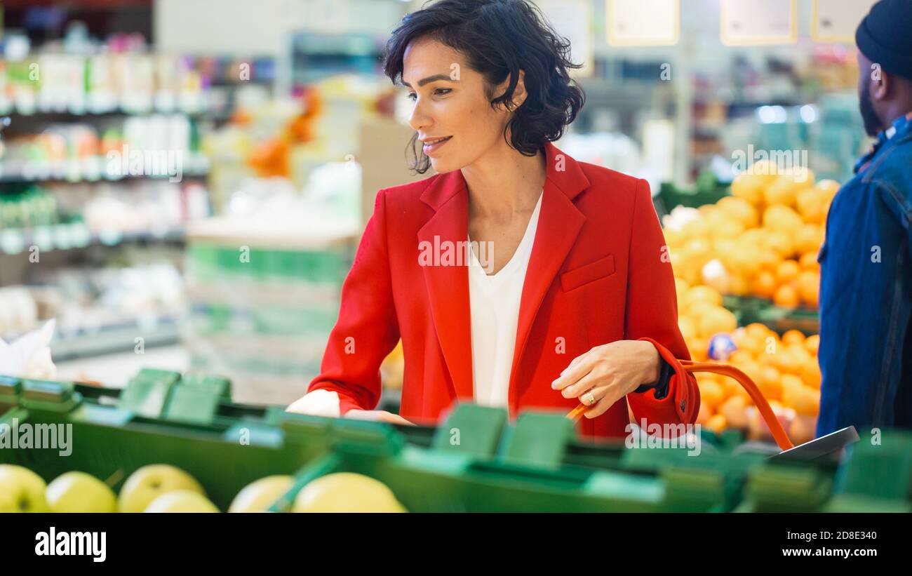 At the Supermarket: Portrait of the Beautiful Smiling Woman Choosing Organic Fruits In the Fresh Produce Aisle and Puts them into Shopping Basket. Stock Photo