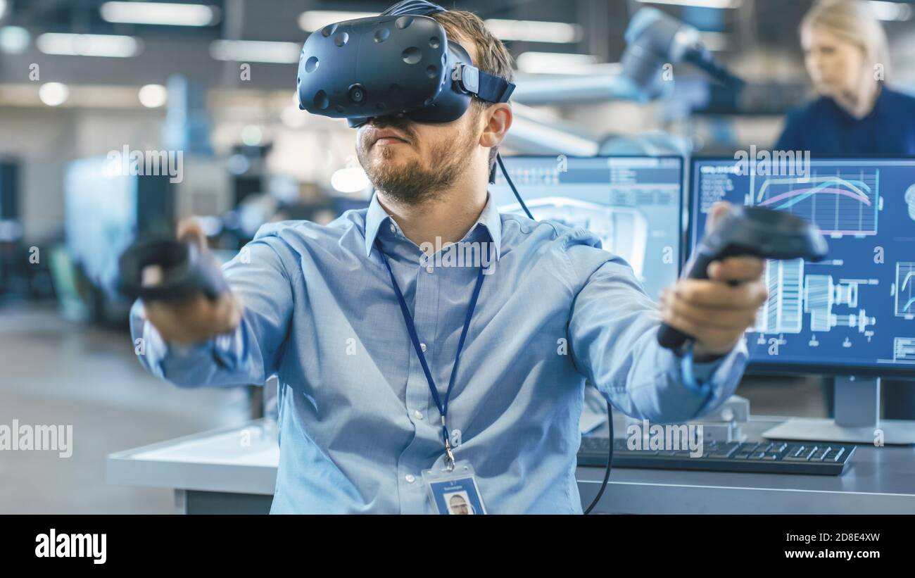 Portrait Shot of the Industrial Engineer Wearing Virtual Reality Headset and Using Controllers, ready to Work. In the Background Manufacturing Plant Stock Photo