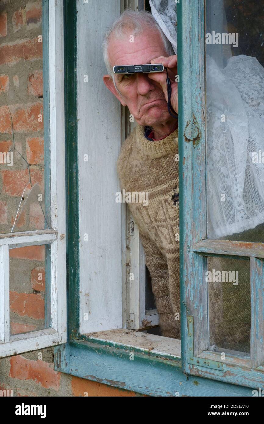 miserable looking older male curtain twitcher spying on neighbours from window to find anyone breaking coronavirus pandemic lockdown rules Stock Photo