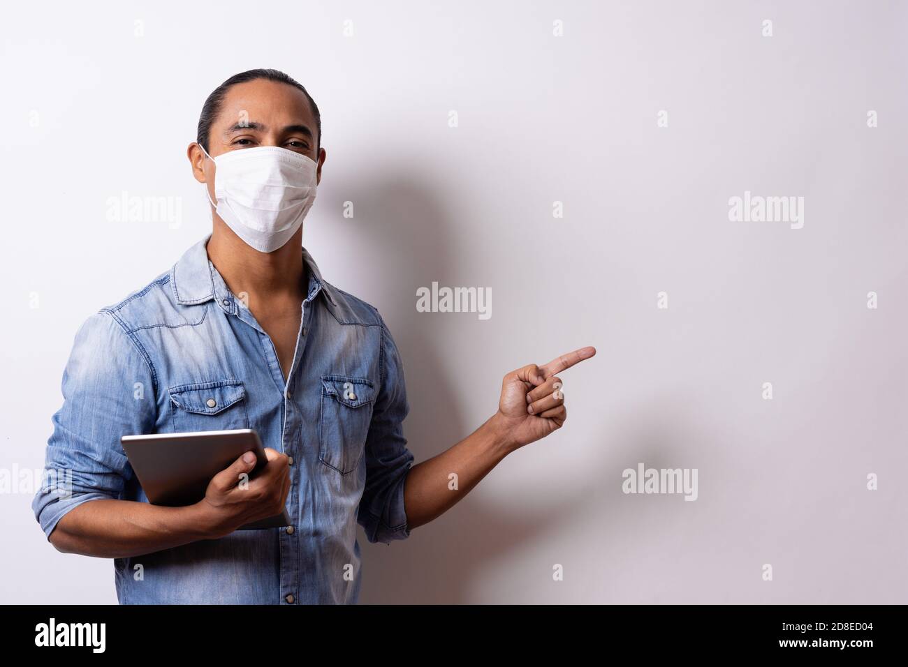 Latino man with facial mask holds a tablet and points to the empty space on white background. Social distance. Stock Photo