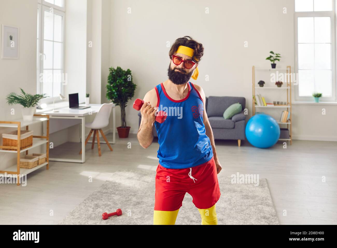 Bearded funny man in bright sport clothes doing workout with dumbbels at home Stock Photo