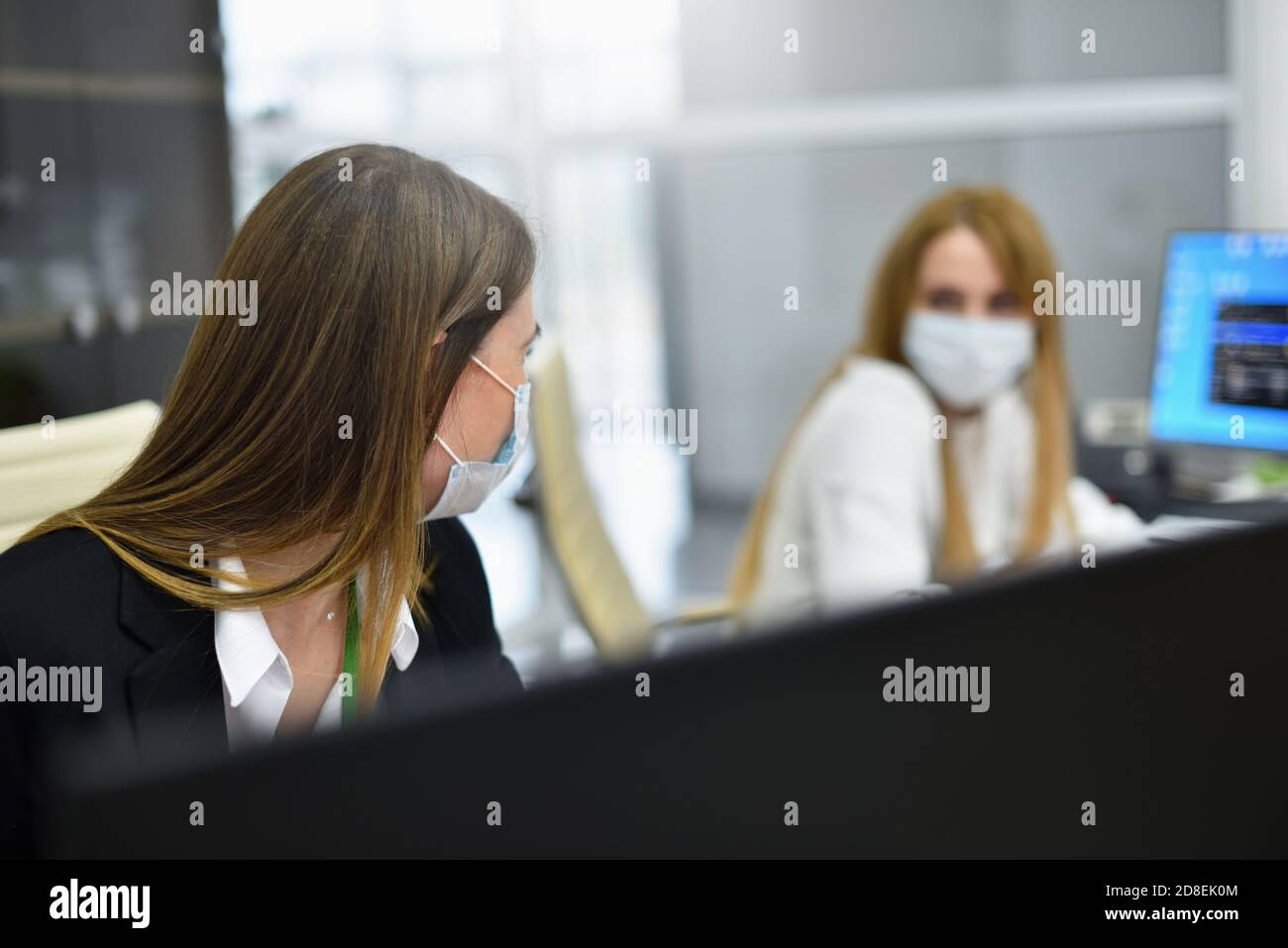 Two female employees office managers in face masks talking at workplace in modern office Stock Photo