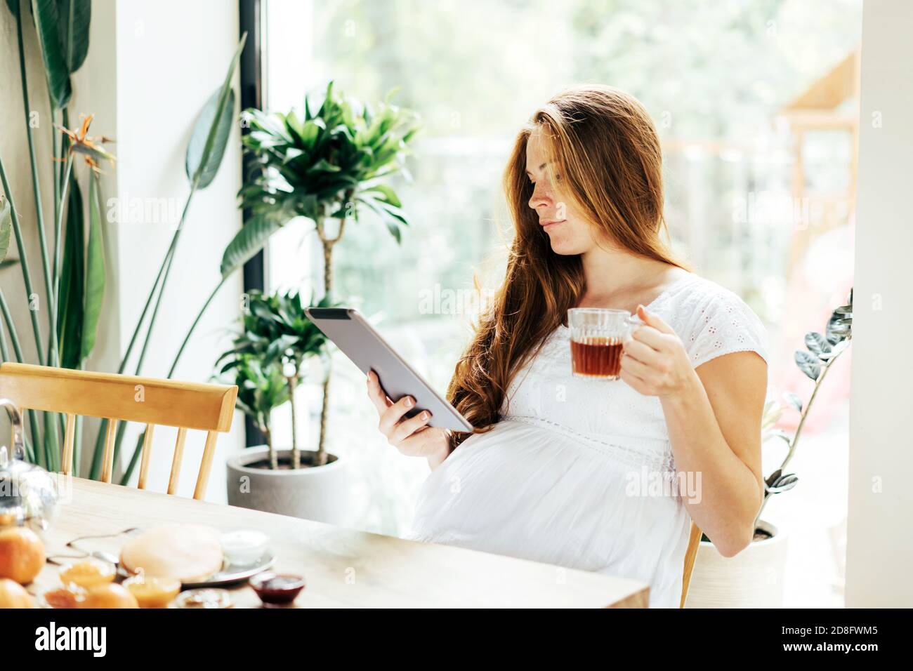 Young modern pregnant woman working on a tablet at breakfast. Stock Photo