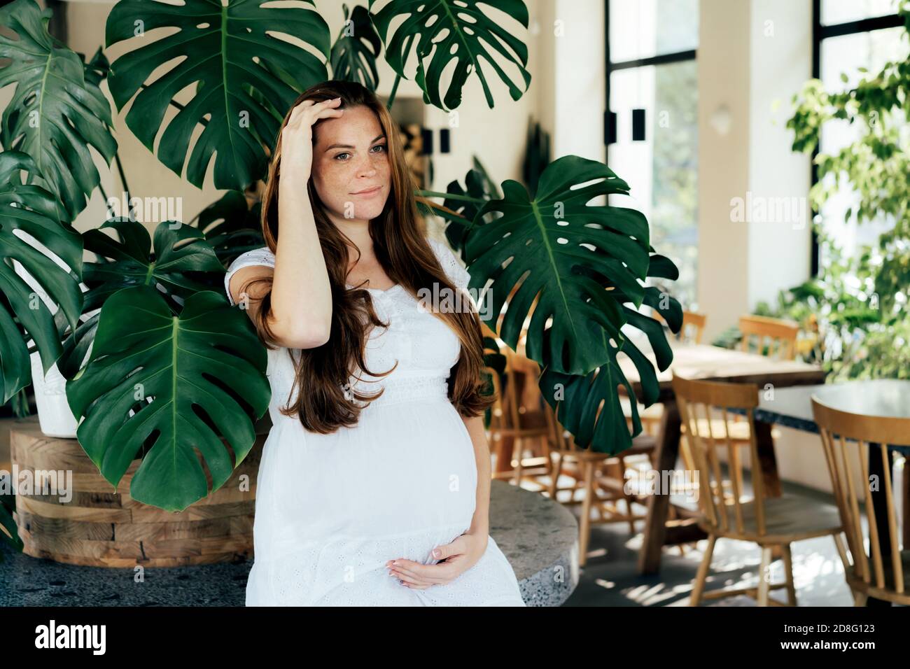 Nice young redhead pregnant woman in stylish interior with tropical monsters. Stock Photo