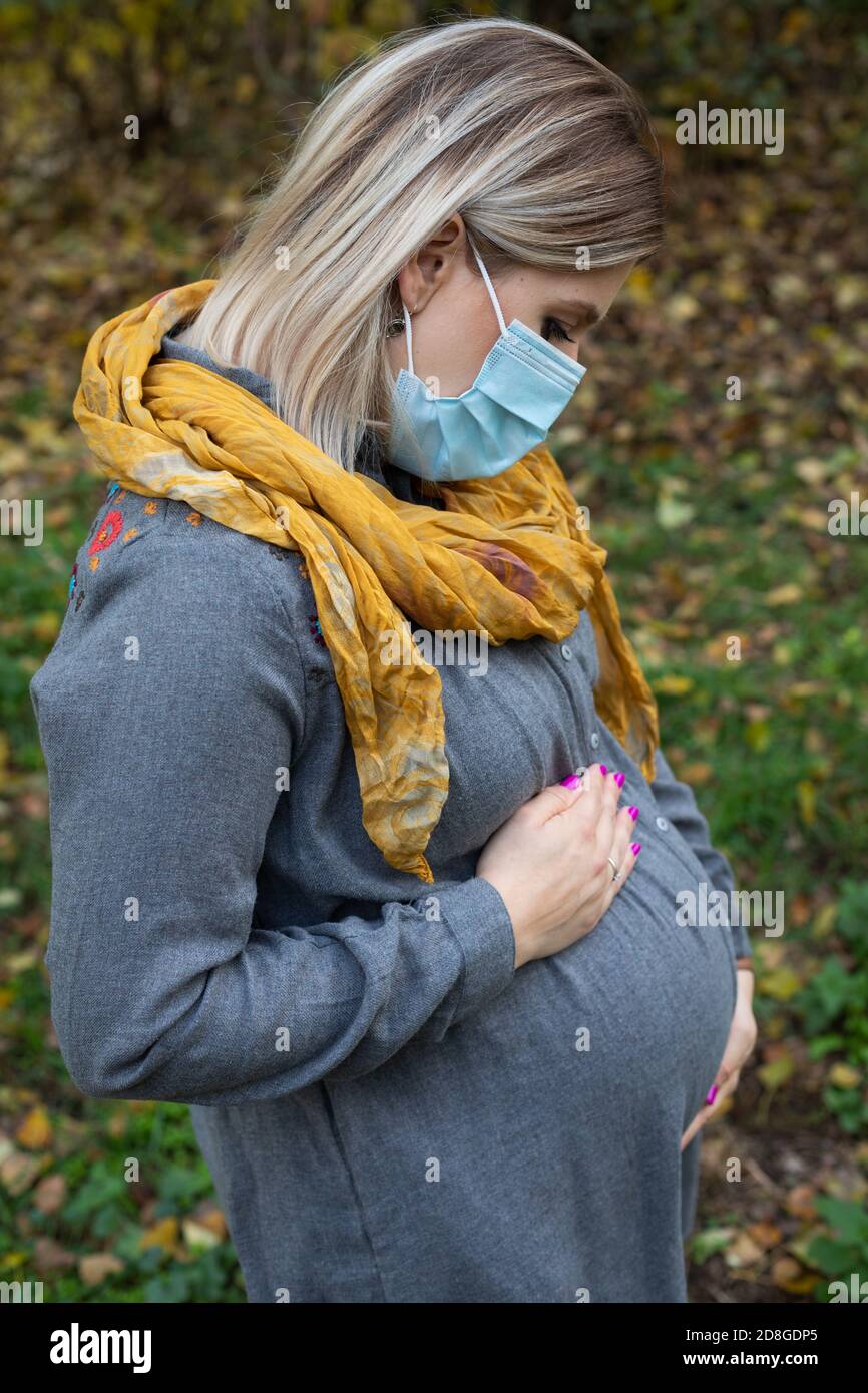 Beautiful pregnant woman in the park wearing surgical mask because of covid-19 global pandemic. Third trimester baby bump Stock Photo