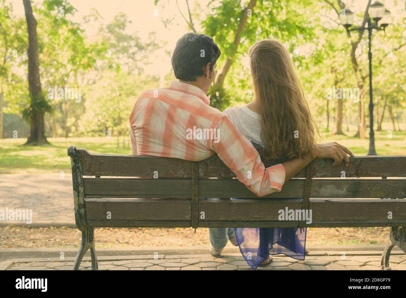 Back view of multi ethnic couple sitting on wooden bench in love while enjoying the scenic view of peaceful green park Stock Photo