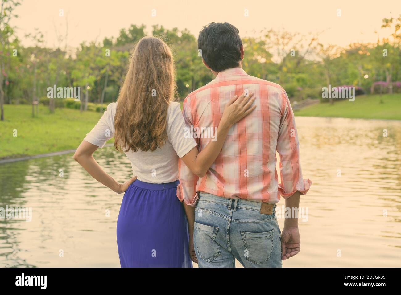Back view of multi ethnic couple standing and enjoying the scenic view of the lake in peaceful green park together Stock Photo