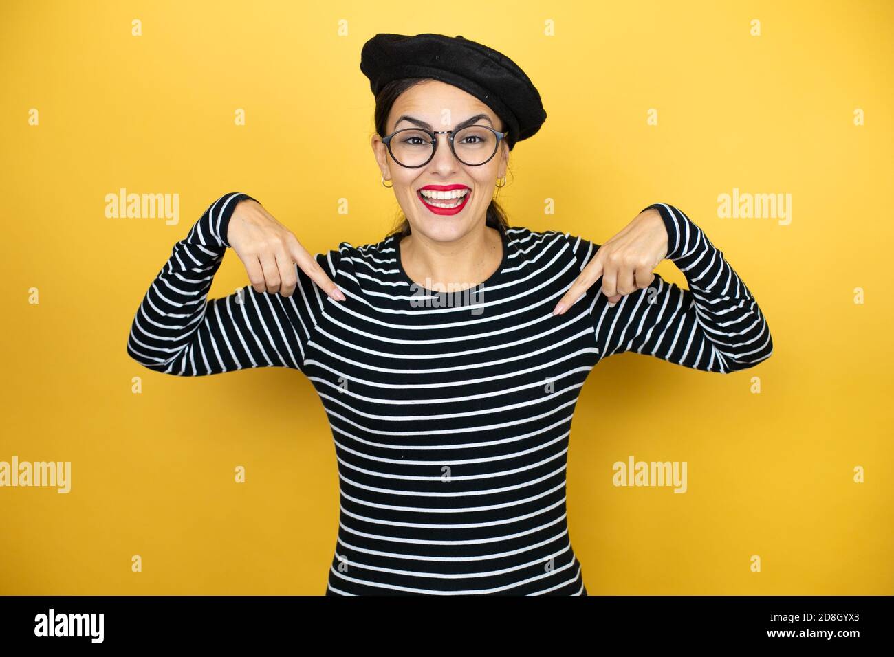 Young beautiful brunette woman wearing french beret and glasses over yellow background looking confident with smile on face, pointing oneself with fin Stock Photo