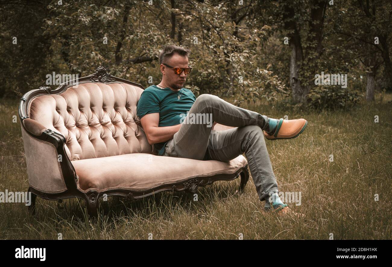 Resting man in grey jeans sitting on a sofa in the middle of garden Stock Photo