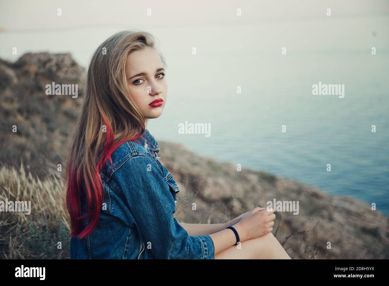 Young blonde girl with bright features smiling on the background of the sea and mountains in a denim skirt and jacket. Background blurred for artistic Stock Photo