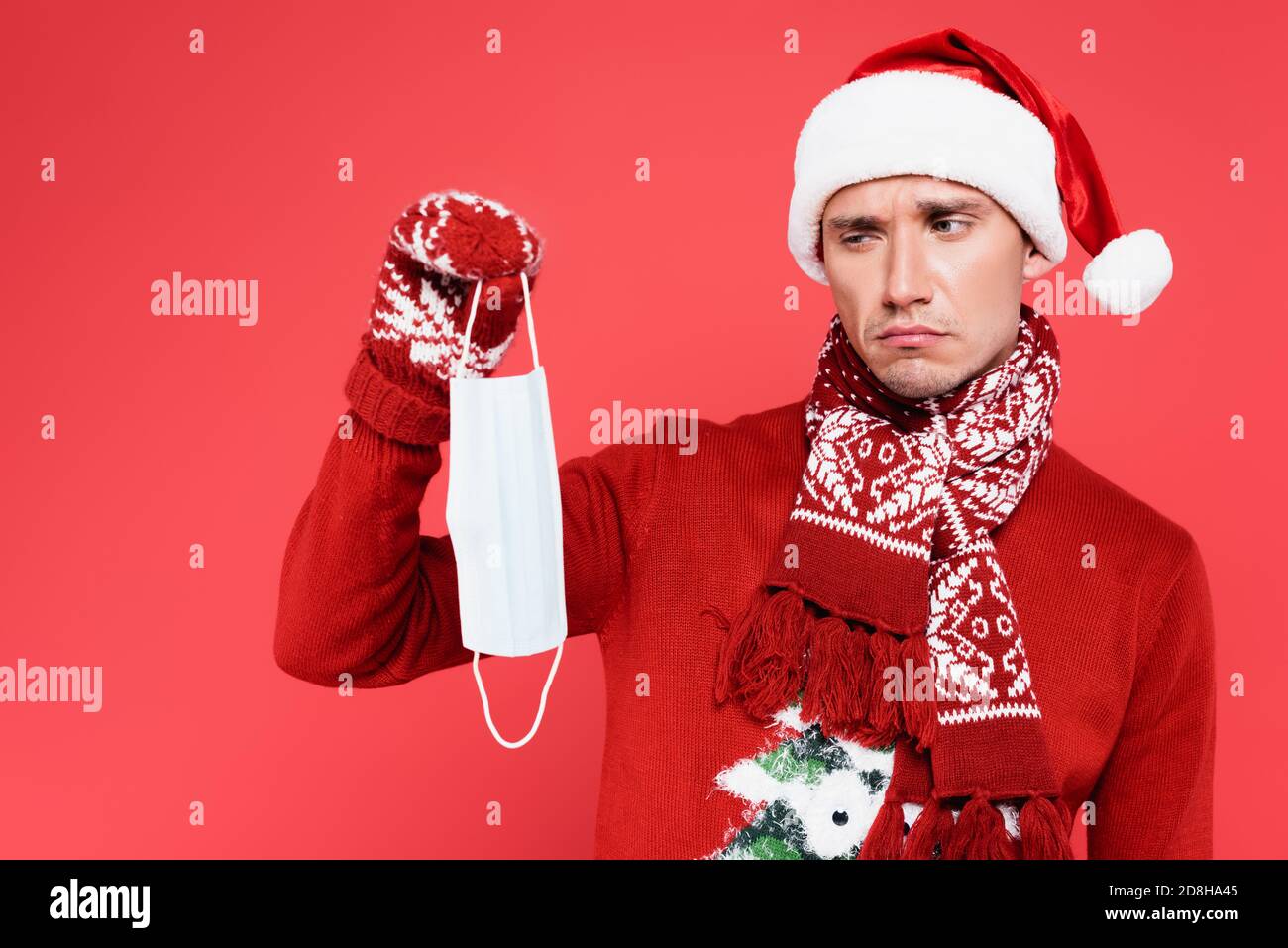 Sad man in santa hat holding medical mask isolated on red Stock Photo