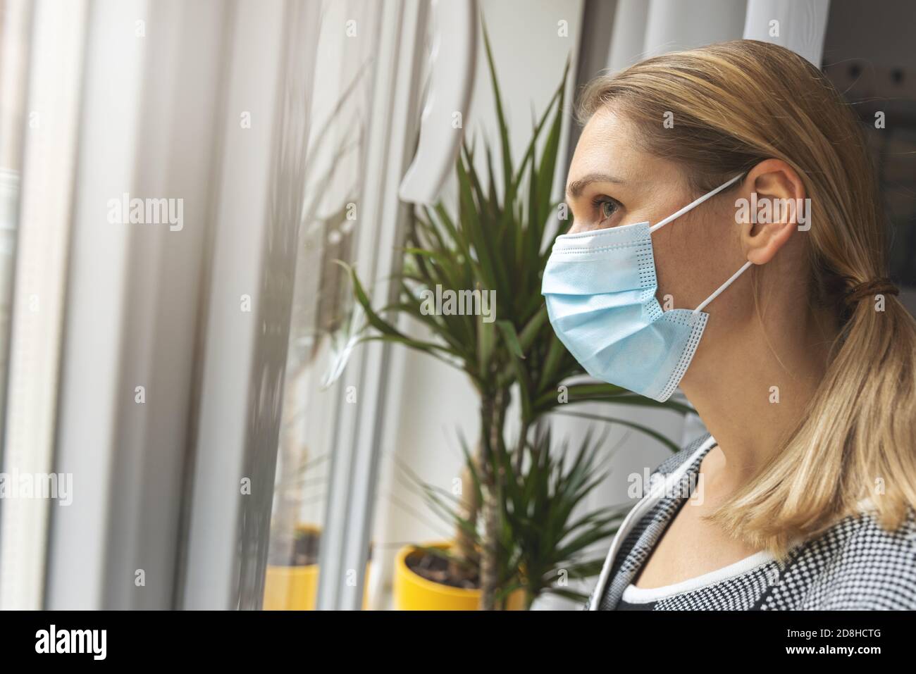 home quarantine - young depressed woman with medical face mask looking through window Stock Photo