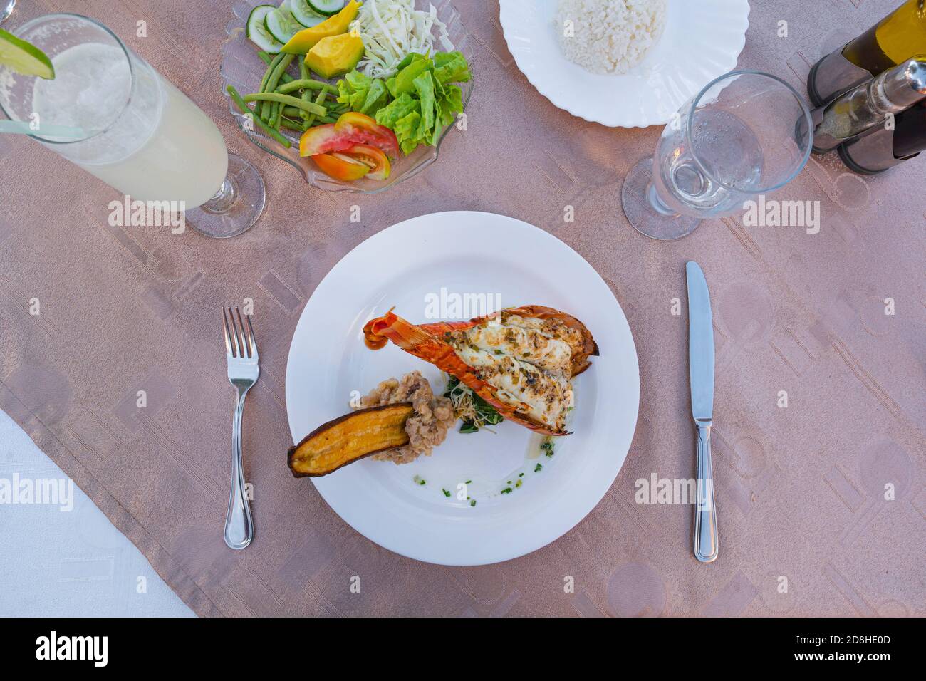 Grilled lobster is beautifully placed on a plate along with side dish of mashed potatoes, fried banana, vegetables and rice and a glass water Stock Photo