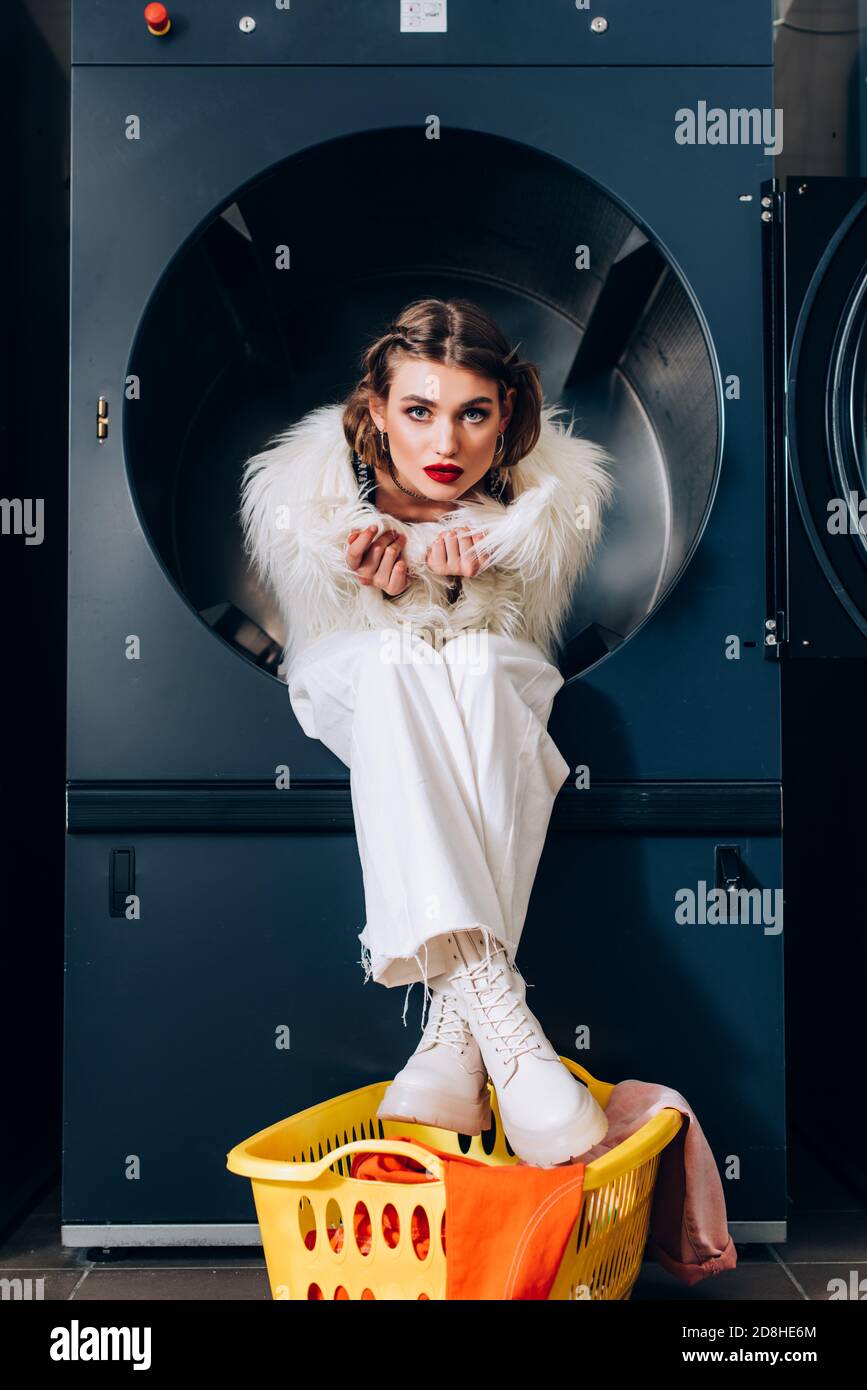 trendy woman in white faux fur jacket sitting in washing machine near