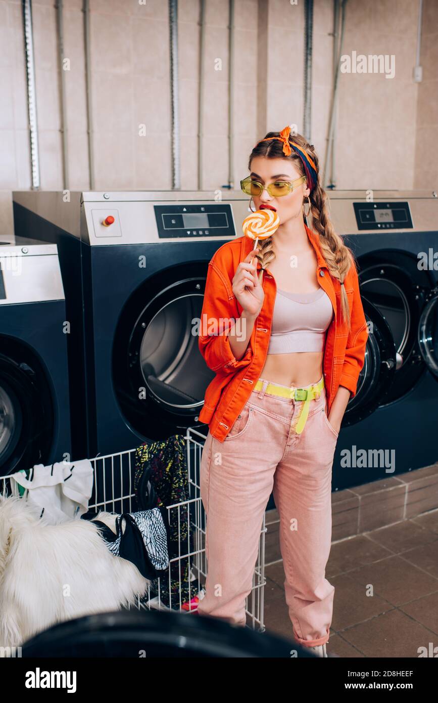 young stylish woman licking lollipop near cart with clothing and washing machines in laundromat Stock Photo
