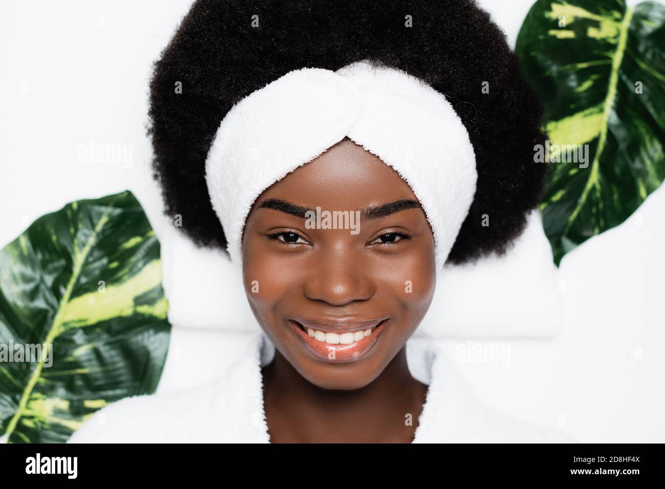 Top view of smiling african american woman with white headband looking at camera, while resting near green leaves in spa salon Stock Photo