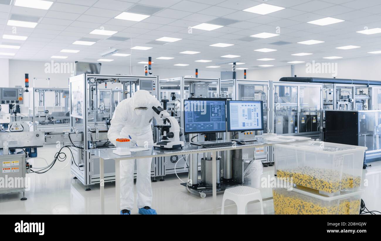 Shot of Sterile High Precision Manufacturing Laboratory where Scientists in Protective Coverall's Use Computers and Microscopes, doing Pharmaceutics Stock Photo