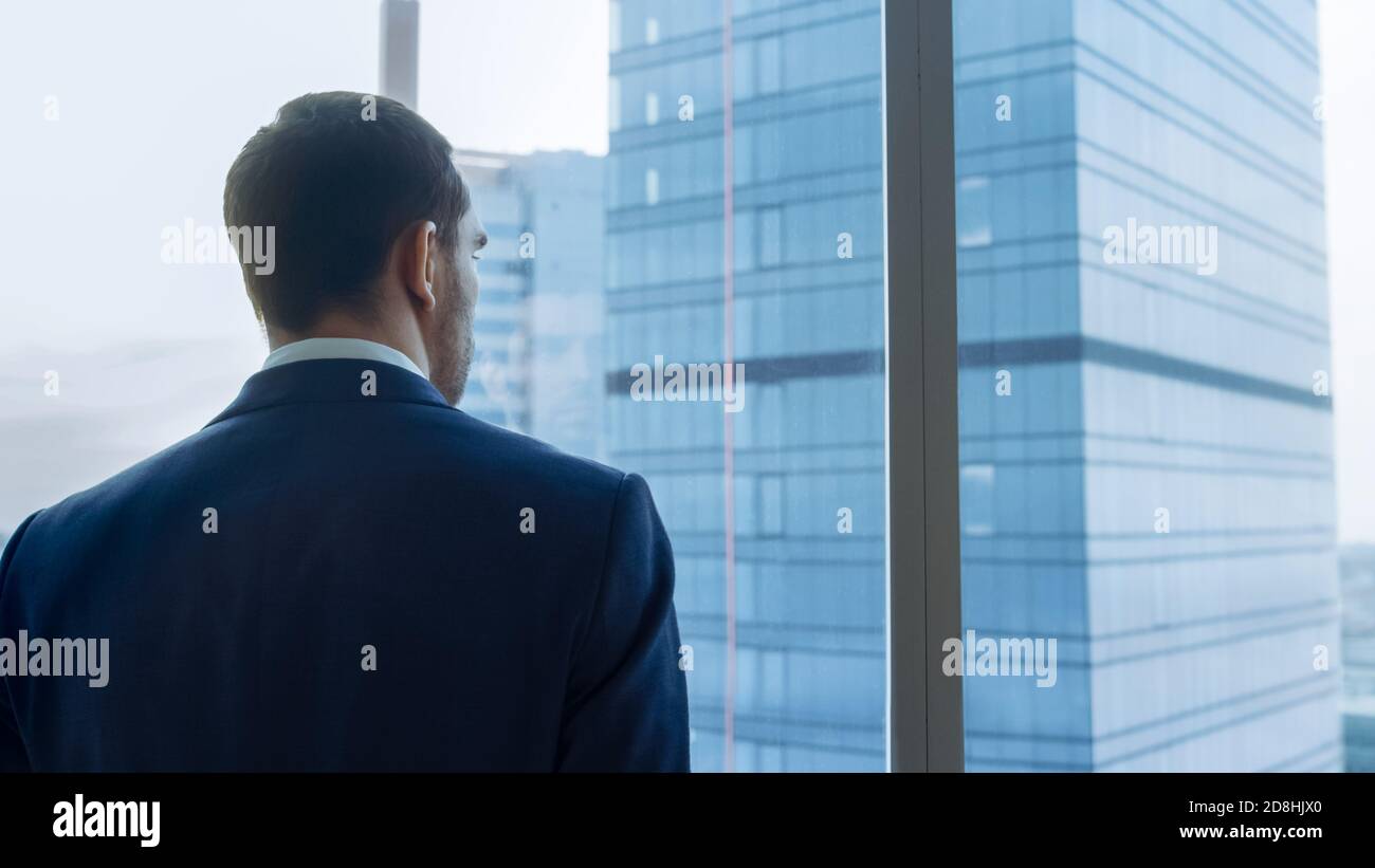 Medium Shot of Successful Businessman wearing a Suit Standing in His Office, Contemplating Next Big Business Deal, Looking out of the Window. Big City Stock Photo