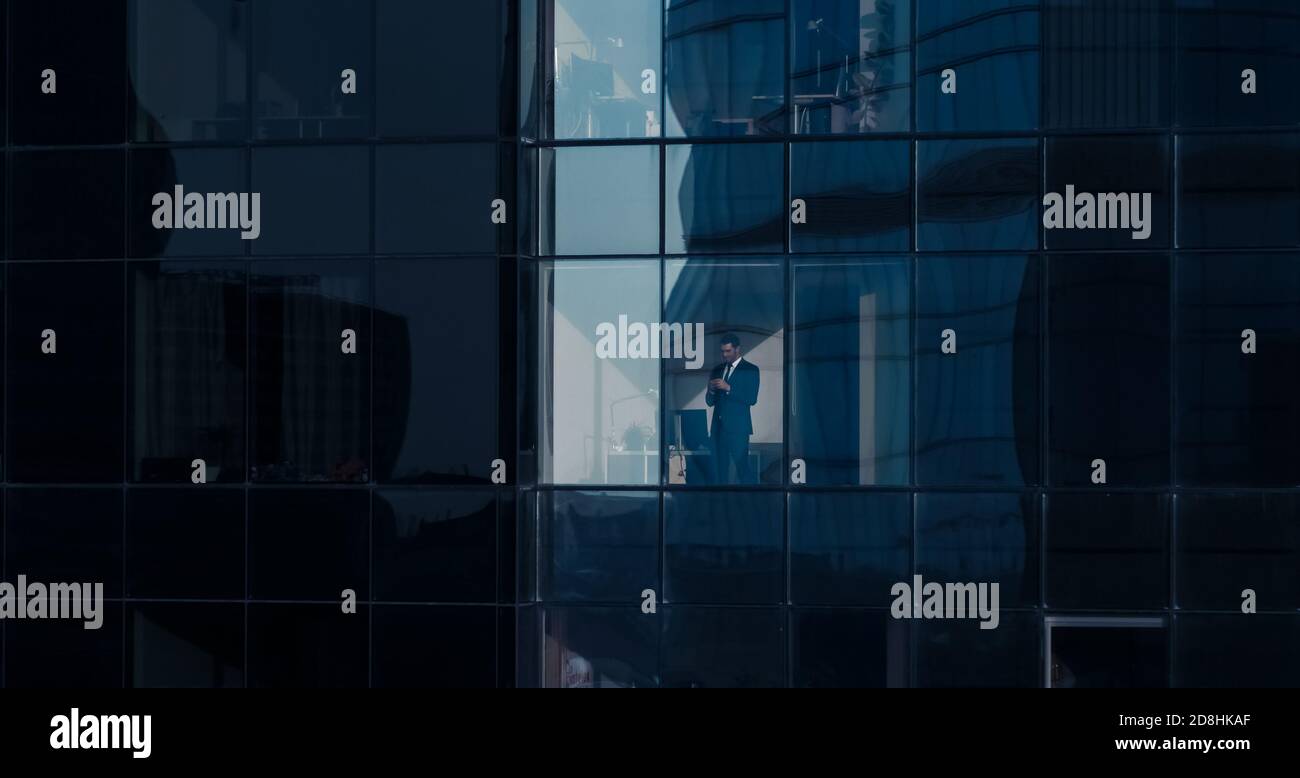 Aerial View Shot: From Outside into Office Building with Businessman Using Mobile Phone and Standing Next to the Office Window. Beautiful Shot of The Stock Photo