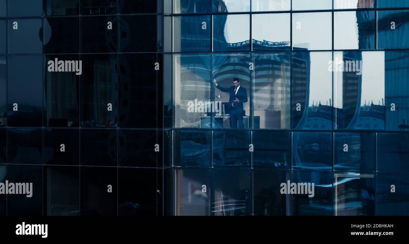 Aerial View Shot: From Outside into Office Building with Businessman Using Mobile Phone and Standing Next to the Office Window. Beautiful Shot of The Stock Photo