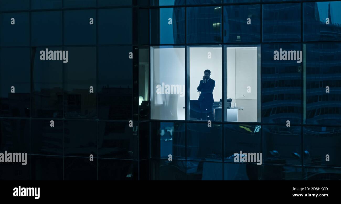 Aerial Shot: From Outside into Office Building with Businessman Using Mobile Phone and Standing by the Office Window. Beautiful Shot of The Financial Stock Photo